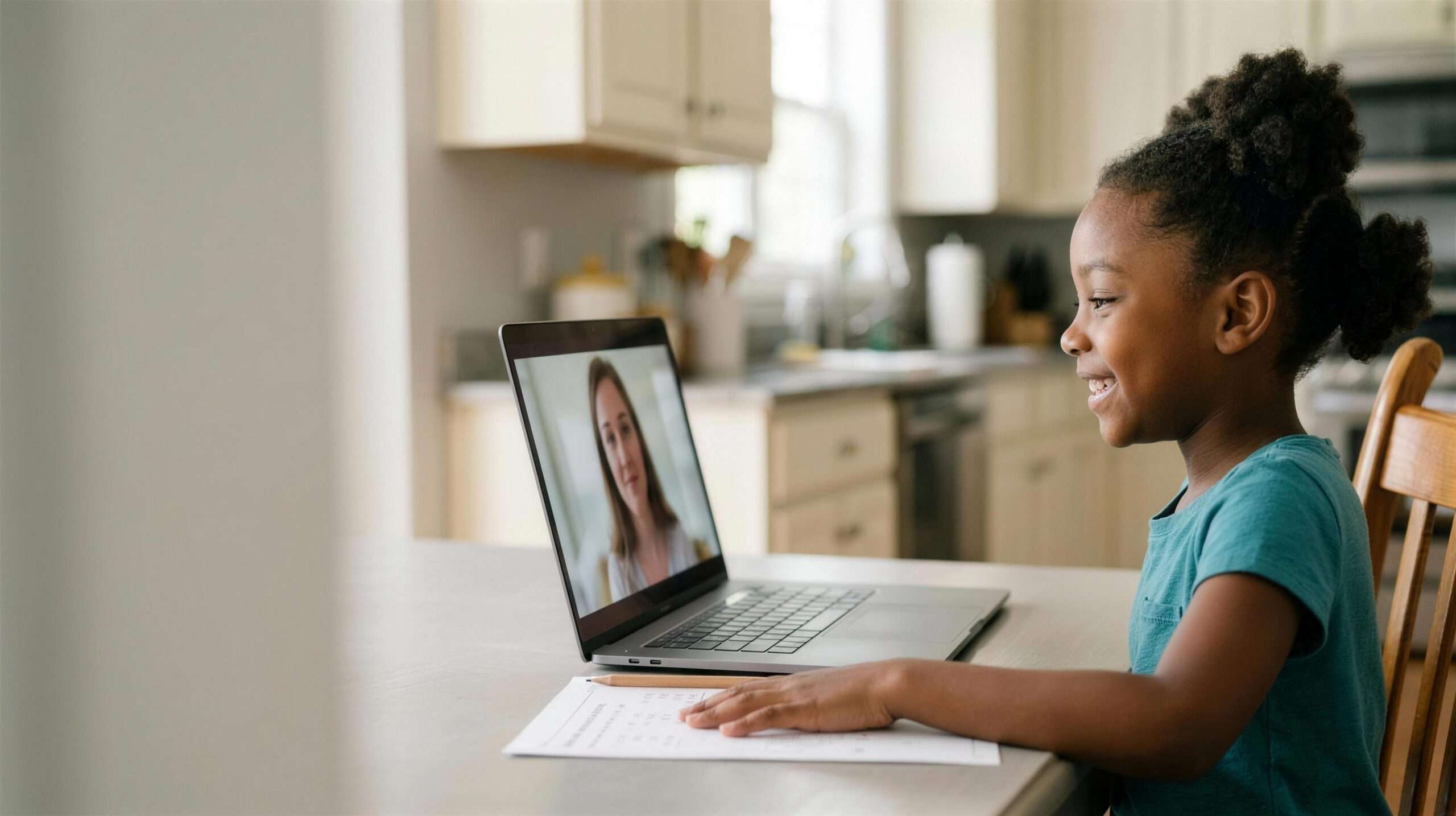 A young elementary student raises a hand excitedly during an online one-on-one tutoring session at the kitchen table.
